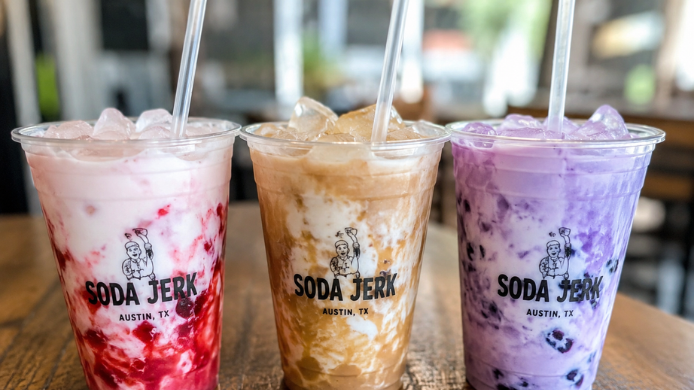 Tres vasos de dirty soda con hielo y capas cremosas de diferentes sabores, servidos sobre una mesa de madera en una cafetería. Las bebidas muestran colores rosa, caramelo y violeta, reflejando la tendencia viral de mezclar refrescos con crema y siropes.