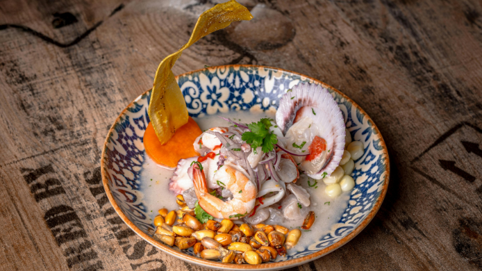 Ceviche peruano con pescado y marisco, cebolla roja, cilantro, maíz tostado y plátano crujiente, servido en un cuenco decorado.