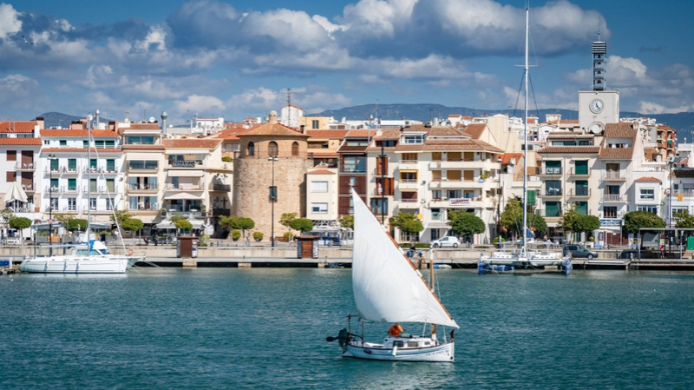 Vista del puerto de Cambrils, Tarragona, con un velero navegando frente al paseo marítimo, la Torre del Puerto y los edificios del casco urbano bajo un cielo azul con nubes.