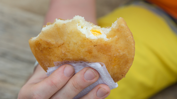 Primer plano de una arepa de huevo tradicional colombiana sostenida con la mano, mostrando su característico relleno de huevo cocido y su masa dorada y crujiente recién frita.