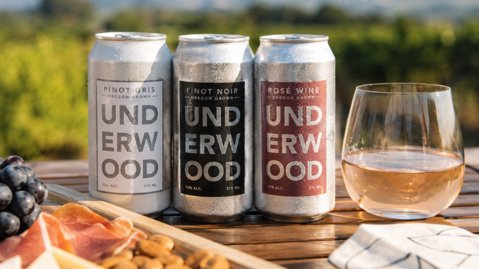 Latas de vino blanco, tinto y rosado servidas junto a una copa y una tabla de aperitivos, con un viñedo al fondo.