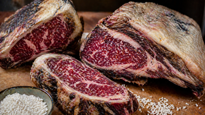 Cortes de carne madurada con textura visible y marmoleado sobre tabla de madera junto a granos de koji en primer plano