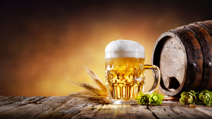 Jarra de cerveza rubia con espuma junto a un barril de madera, espigas de cebada y lúpulo sobre una mesa rústica.