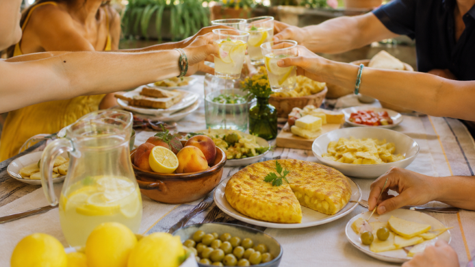 Mesa de verano al aire libre con tortilla española, limonada, fruta amarilla y varias personas brindando durante una comida compartida, una imagen que evoca la felicidad asociada al Yellow Day.