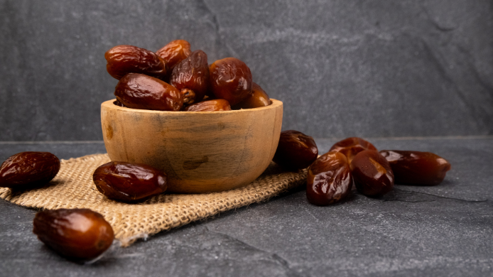 Cuenco de madera con dátiles maduros sobre una superficie de piedra oscura, mostrando distintas piezas de esta fruta tradicional apreciada por su dulzor natural y su valor gastronómico.
