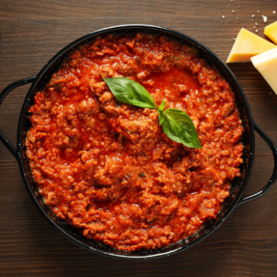 Salsa boloñesa tradicional en cazuela negra, decorada con hojas de albahaca y acompañada de queso sobre una mesa de madera.
