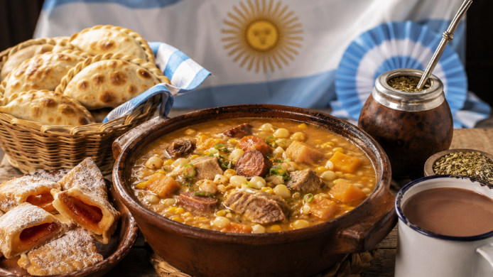Mesa con platos tradicionales argentinos para celebrar el Día de la Patria, con locro, empanadas, pastelitos, mate y chocolate caliente junto a la bandera argentina.