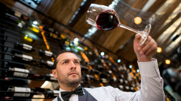 Sumiller observando una copa de vino tinto durante una cata en una bodega con botellas al fondo