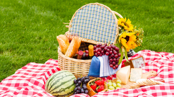 Cesta de picnic sobre una manta de cuadros en el césped con frutas frescas, pan, queso, flores y una botella de vino, símbolo de la nueva tendencia gastronómica de comer al aire libre.
