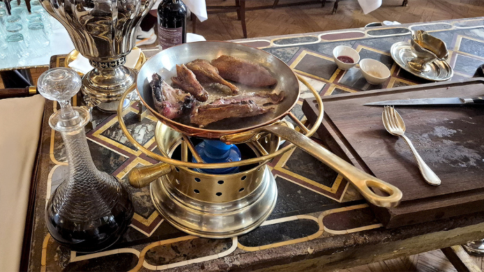 Servicio de pato a la prensa en una sartén de cobre sobre una mesa de restaurante, acompañado de una jarra de salsa y cubertería.