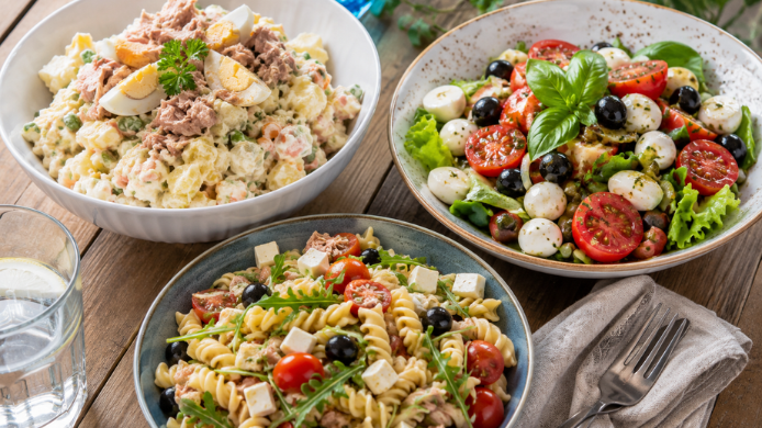 Tres ensaladas de verano servidas sobre una mesa de madera al aire libre: una ensaladilla rusa tradicional, una ensalada mediterránea con mozzarella y tomates cherry y una ensalada de pasta, ideales para los días más calurosos del año.