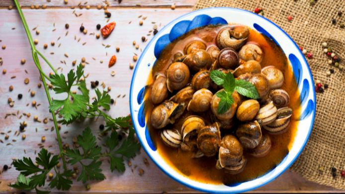 Plato de caracoles en salsa tradicional servidos en cuenco de cerámica, acompañados de hierbas aromáticas y especias, representación de una receta clásica de la gastronomía española.