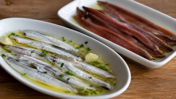 Boquerones en vinagre y anchoas en aceite servidos en platos blancos sobre una mesa de madera
