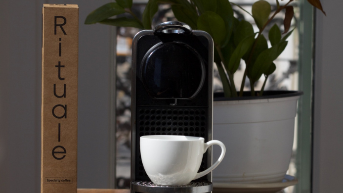Caja de café Rituale junto a una cafetera de cápsulas y una taza recién preparada, imagen que representa la apuesta por el café de especialidad en cápsulas compostables y sostenibles.