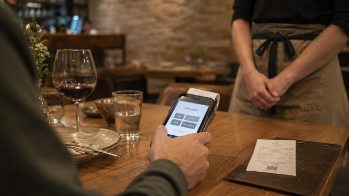 Comensal decide si añadir propina en un datáfono al finalizar la comida mientras el personal de sala espera junto a la mesa.