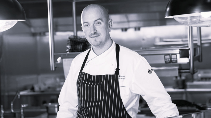 Retrato en blanco y negro del chef Luca Cesarini en una cocina profesional, vestido con chaquetilla y delantal de rayas.