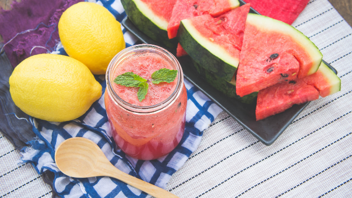 Un tarro de cristal con batido de sandía, cubierto con hojas de menta fresca y un limón entero, se sienta en una mesa de madera. Sandia-recetas.jpg muestra el batido y los ingredientes.
