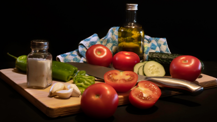 Ingredientes tradicionales para preparar gazpacho casero (tomates, pimiento, pepino, ajo, sal y aceite) dispuestos en una tabla de madera con un cuchillo.