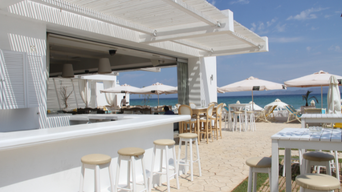 Un chiringuito de playa con bar exterior blanco, sombrillas y vista al océano en un día soleado.