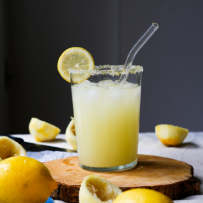 Vaso de limonada casera con hielo, pajita de cristal y borde escarchado, rodeado de limones enteros y exprimidos.