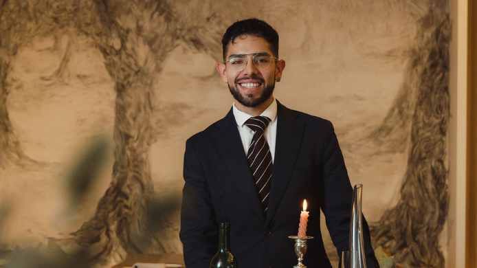 Brandon Jordan, vestido con traje oscuro en la sala de Gurisa, junto a una mesa con botella de vino y una vela encendida.