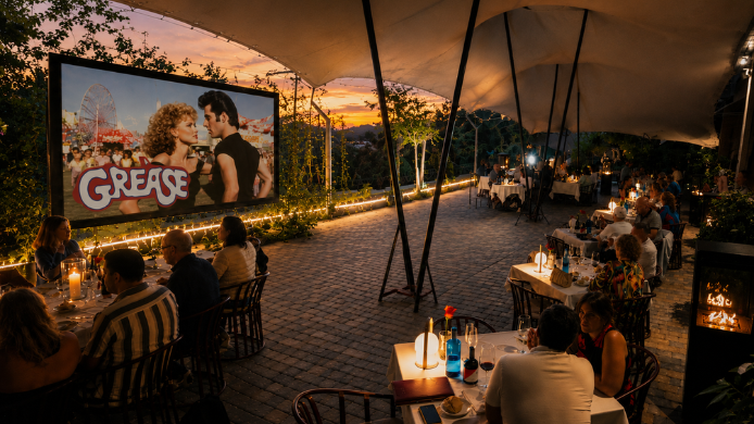 Cena al aire libre en una terraza entre viñedos durante la proyección de Grease, con mesas iluminadas y público disfrutando del cine bajo las estrellas.