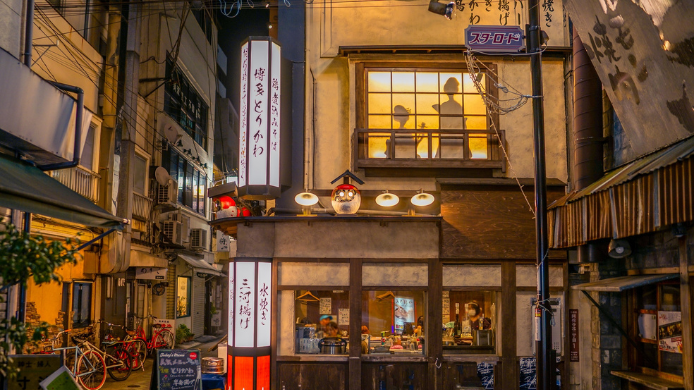 Fachada nocturna de una izakaya japonesa iluminada en una calle estrecha, con faroles, carteles y ambiente urbano.