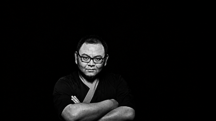 Retrato en blanco y negro del chef Xiao Wang con gafas y los brazos cruzados sobre un fondo negro.