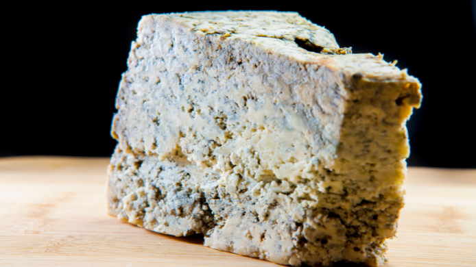 Pieza de queso Cabrales con vetas azul verdosas sobre una tabla de madera y fondo negro.