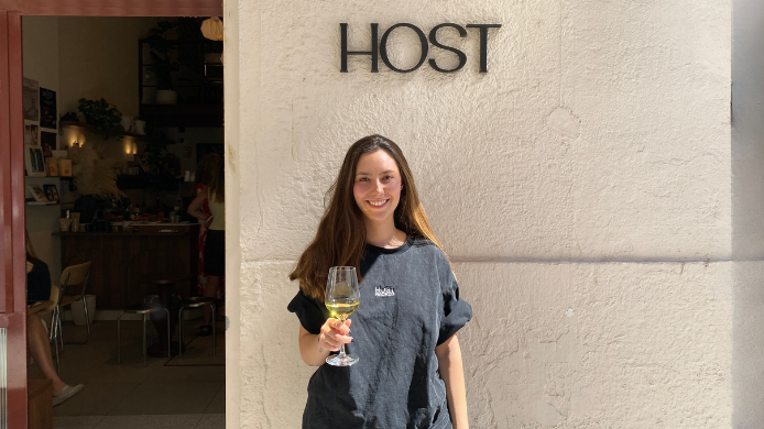 Mariana Faccini en la entrada de Host Madrid sosteniendo una copa de vino blanco junto al rótulo del local