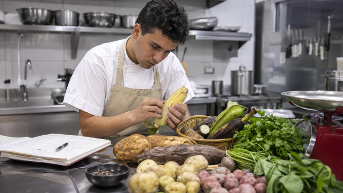 Joven chef colombiano examina una mazorca junto a tubérculos, plátanos y hierbas mientras toma notas en una cocina profesional.