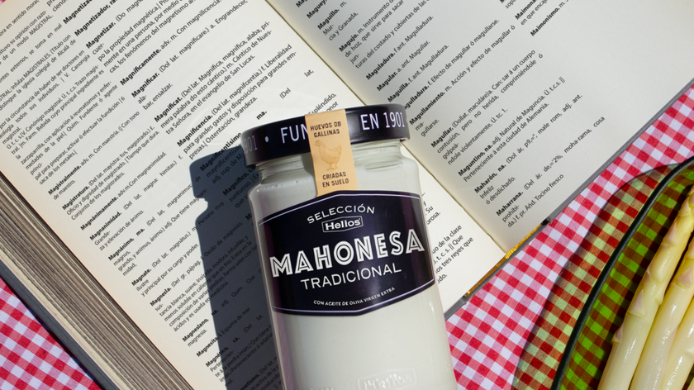 Bote de mahonesa tradicional Helios sobre un libro abierto y mantel de cuadros junto a espárragos blancos