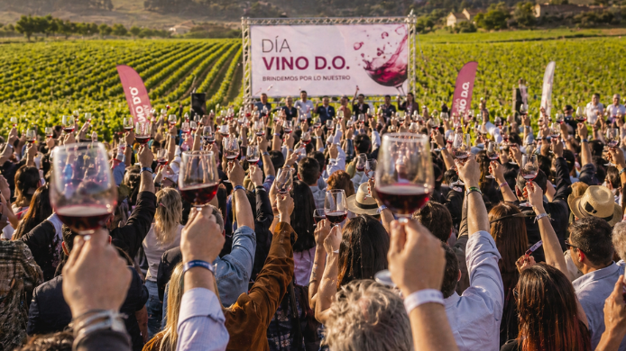 Multitud brindando con copas de vino en un viñedo durante el Día Vino D.O., con escenario al fondo y paisaje de viñas en España.