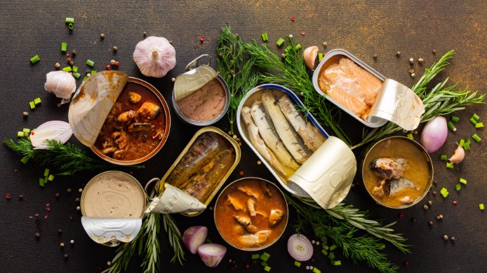 Selección de conservas de pescado abiertas con sardinas, salmón, atún, mejillones y otros productos en aceite, acompañadas de hierbas aromáticas, ajo y especias, una imagen que ilustra el aprovechamiento del aceite de las latas en la cocina.