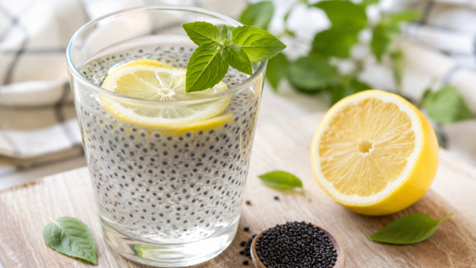 Vaso de agua con semillas de albahaca hidratadas y rodajas de limón sobre una mesa de madera, acompañado de hojas frescas de albahaca y semillas secas, una bebida rica en fibra soluble asociada a una mayor sensación de saciedad.