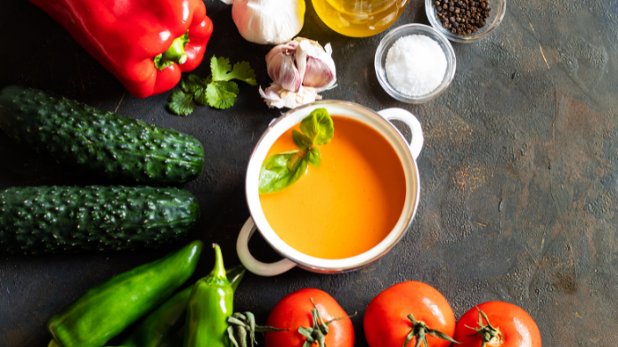 Plato de gazpacho casero rodeado de hortalizas frescas como tomates, pepinos y pimientos sobre una mesa oscura.