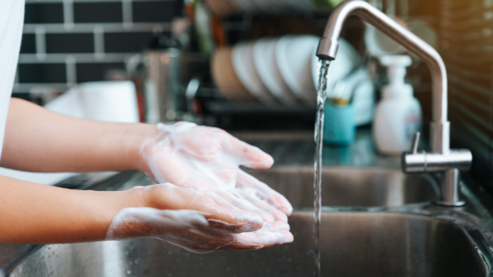 manos frotándose enérgicamente con espuma de jabón bajo el agua de un grifo abierto en una cocina. La imagen resalta la importancia del lavado de manos para prevenir infecciones alimentarias como la salmonelosis.