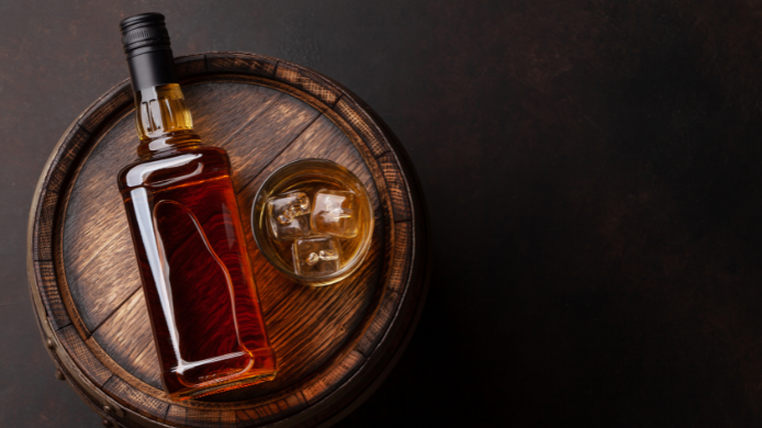 Botella de whisky y vaso con hielo sobre una barrica de madera, vistos desde arriba sobre un fondo oscuro.