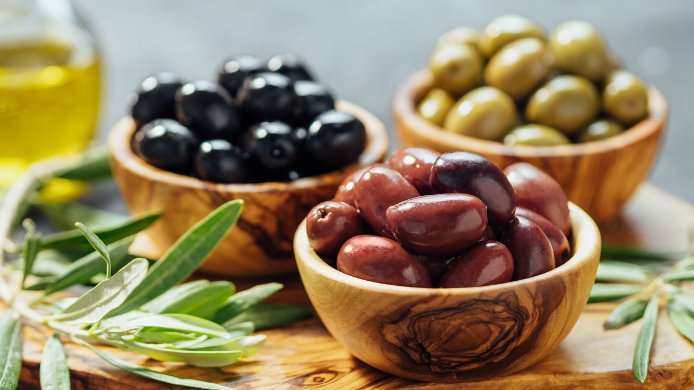 Cuencos de madera con distintas variedades de aceitunas verdes, negras y moradas sobre una tabla rústica, acompañadas de ramas de olivo y aceite de oliva.