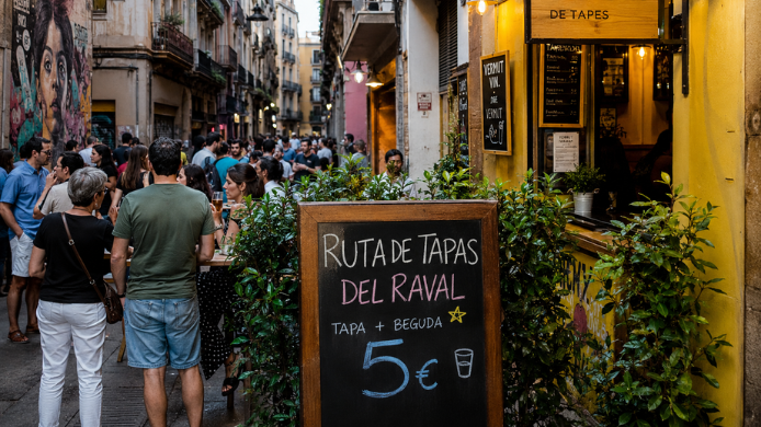Ambiente de tapas en una calle del Raval de Barcelona durante la Festa Major, con bares, vecinos y una pizarra de ruta gastronómica.