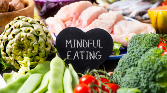 na pequeña pizarra negra con forma de corazón que tiene escrito "MINDFUL EATING", rodeada de alimentos saludables como verduras frescas y carne de pollo cruda