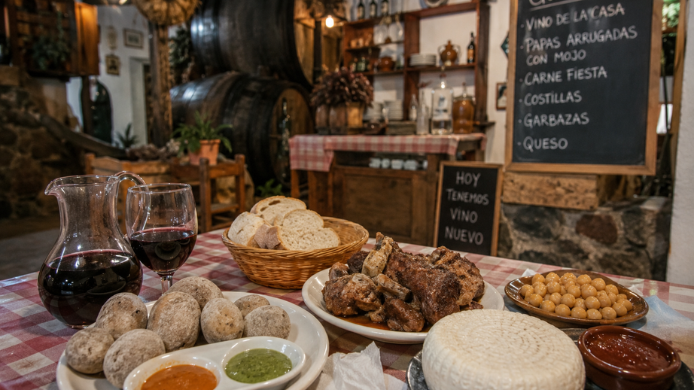 Mesa de un guachinche tradicional de Tenerife con vino tinto de cosecha propia, papas arrugadas con mojo, carne, garbanzas, queso canario y pan, en un comedor rústico con barricas de madera.