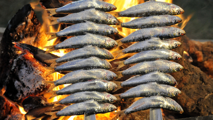 Dos espetos de sardinas ensartadas asándose a fuego abierto sobre leña ardiente. Se ve la piel dorada de las sardinas y las llamas naranjas detrás.